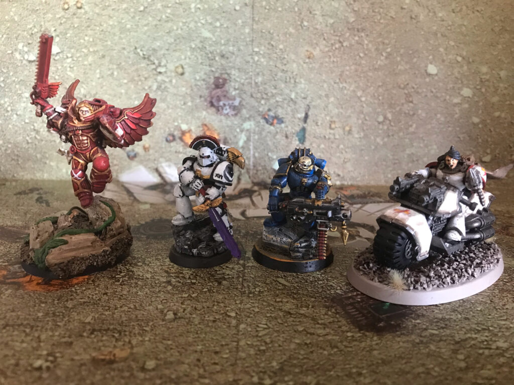 Four models of characters from different Space Marine legions. From left to right, a Blood Angel Apothecary with winged jump pack. A Luna Wolf with a two-handed power sword. A Night Lord with a heavy bolter. A White Scar on a bike.