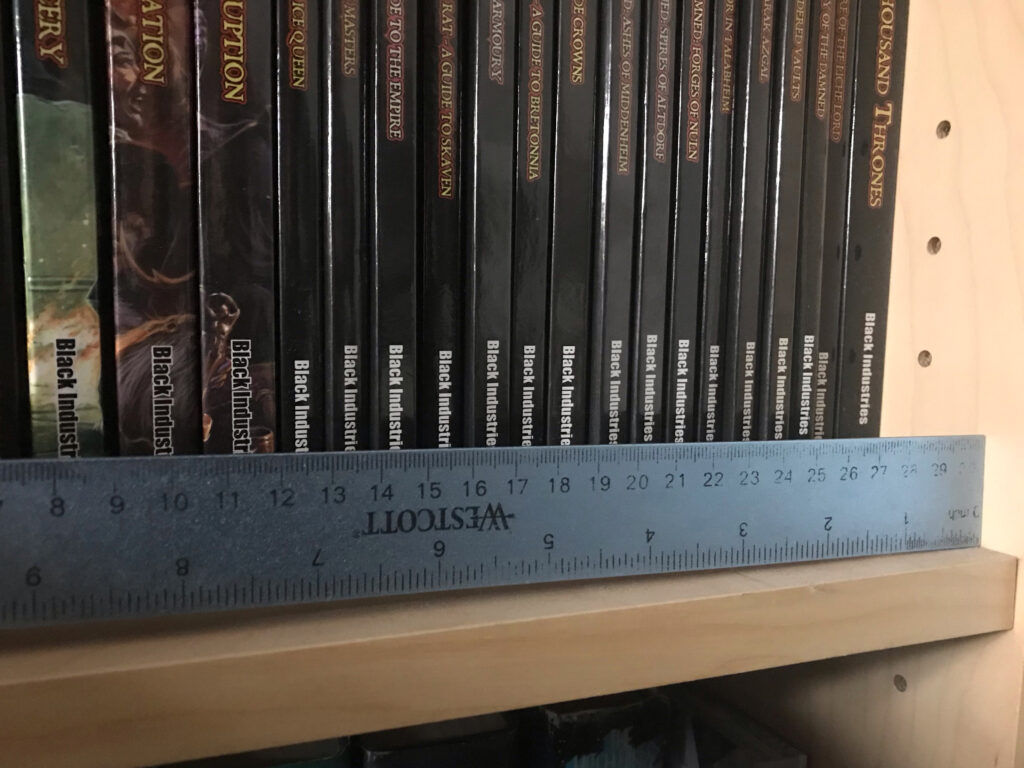 A picture of a shelf of books. The books are all from Warhammer Fantasy Roleplay second edition, published by Black Industries. A metal ruler shows how the shelf is noticeably bending under the weight of the books.