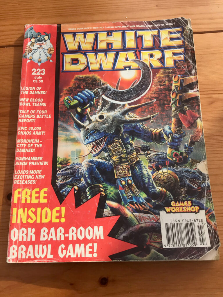 A battered copy of White Dwarf 223. The cover features a Lizardman warrior wearing a stegadon skull helmet, and a list of articles down the left hand side, including Legion of the Damned, New Blood Bowl Teams, and an Ork Bar-Room brawl game