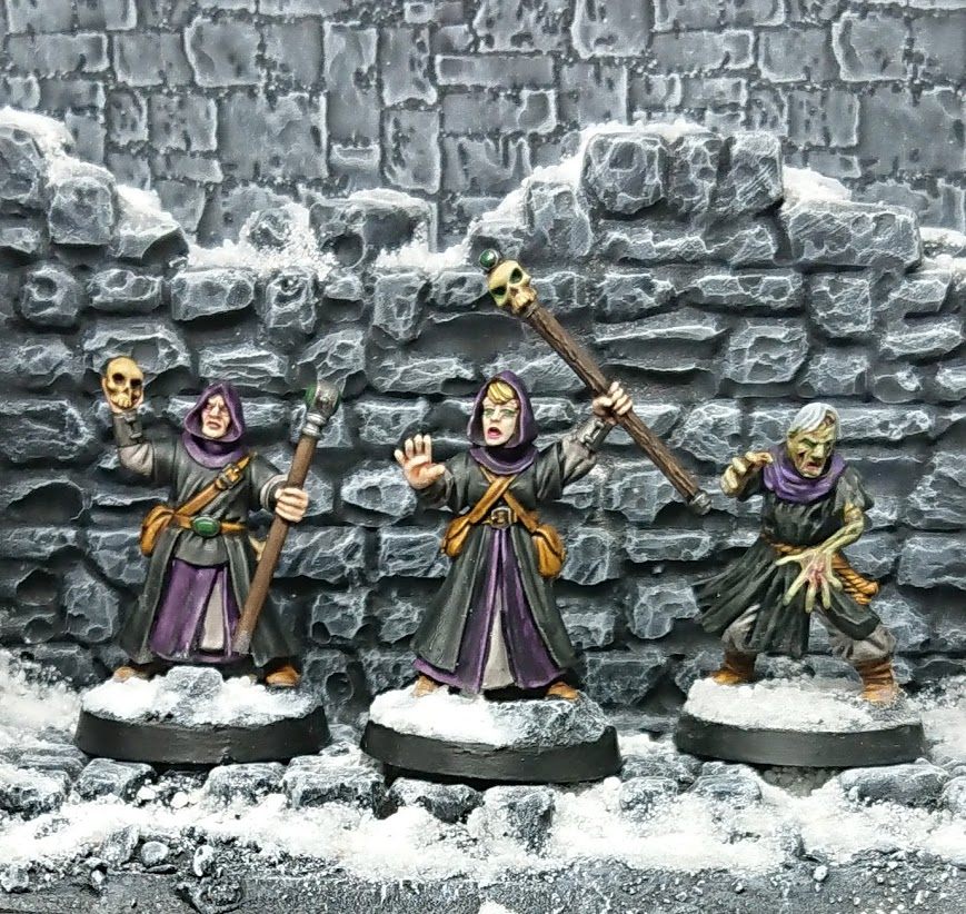 Photograph of a pair of black-robed wizards and a zombie, in front of a background of grey stone and snow.