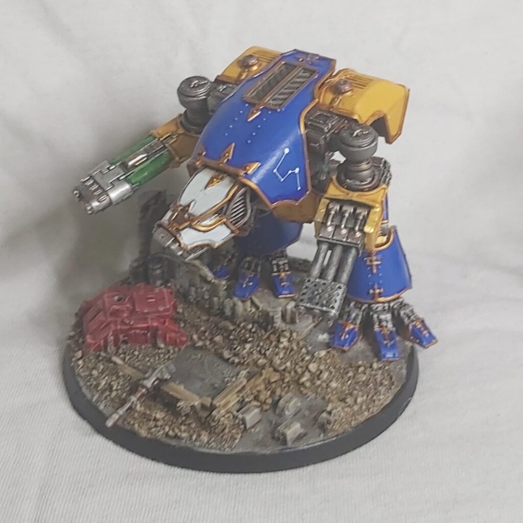 Photograph of a warhound titan painted blue and yellow, mounted on a base decorated to look like a ruined city block
