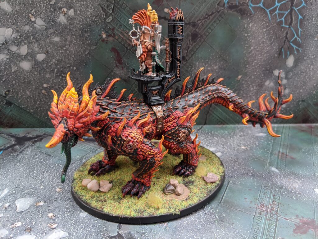 A magmadroth model, painted in a fiery red and black colour scheme on a grassy base.