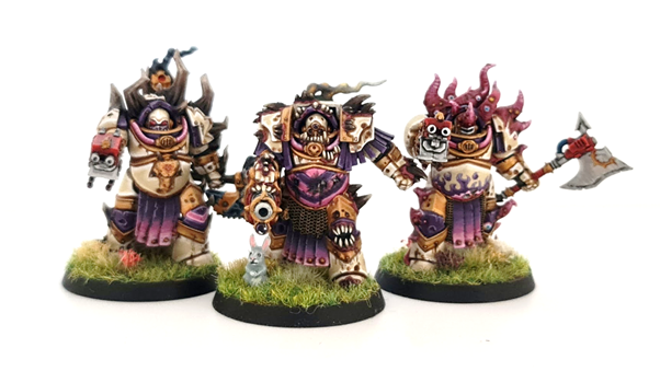 A photo showing the three completed models of the Tainted Cohort. They are painted with pale armour, pink and purple accents and gold trim. Their weapons are accented with red casings. They are mounted on grassy bases with additional flower tufts. A grey rabbit is visible on one base. 
