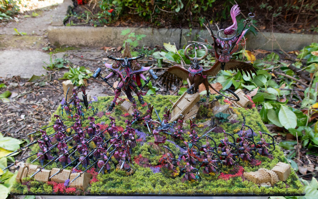 A pink army of elves, on a grassy display board, set outside in a leafy garden
