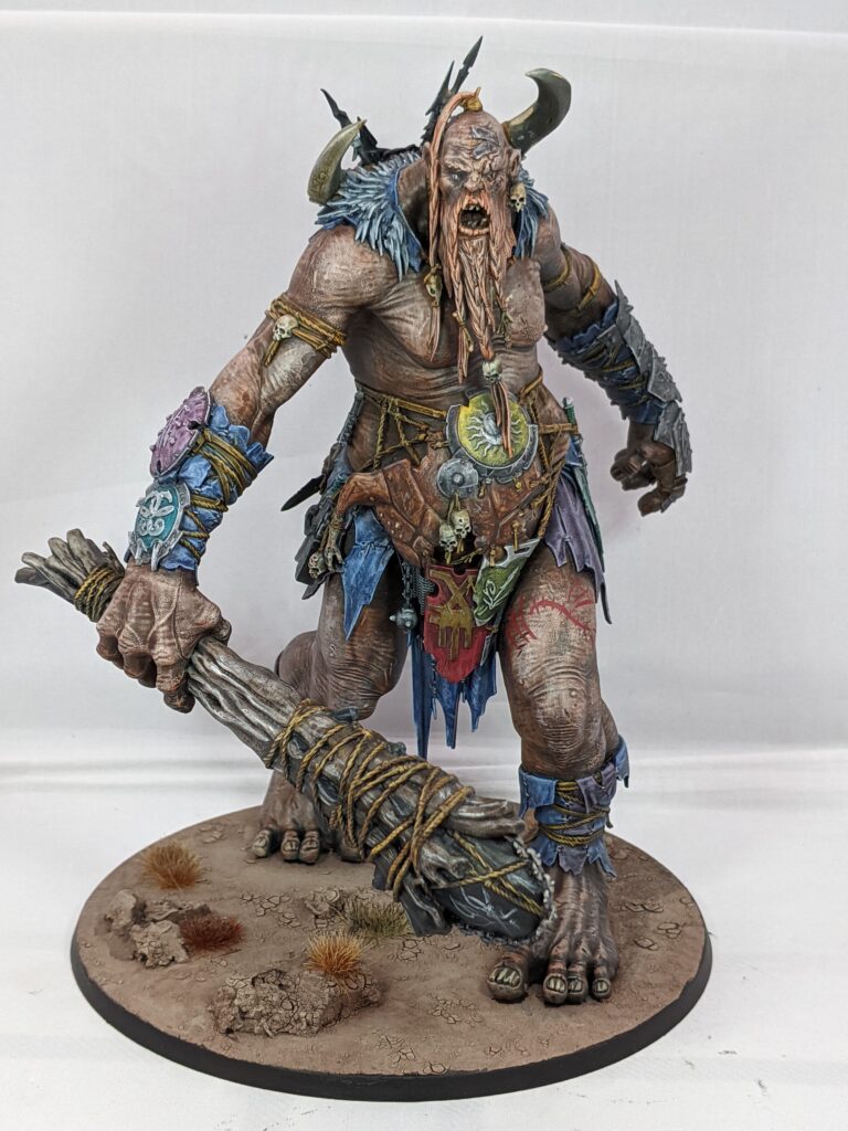 A painted Warstomper Mega-Gargant on a desert themed base. He has a ginger beard and blue and purple clothes. There is a tattoo of a zombie dragon on his left thigh.