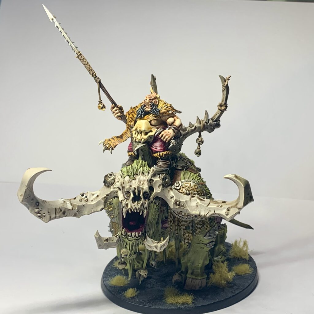 A Mighty Ogor clad in furs, armoured in bone riding atop a moss green beast with towering bone white horns.